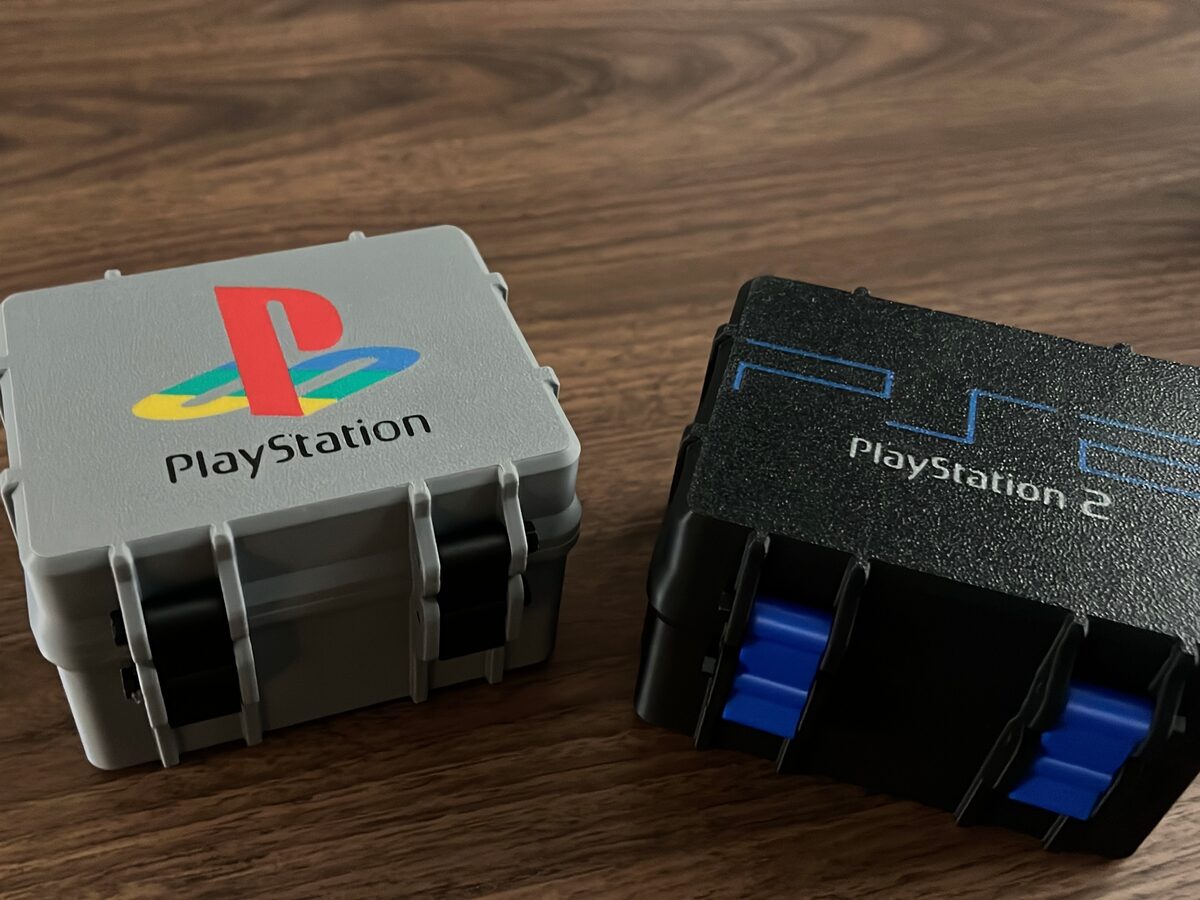 Dos cajas de almacenaje impresas en 3D, una gris y otra negra, con logotipos impresos a varios colores en la tapa y cierres funcionales, sobre una mesa de madera