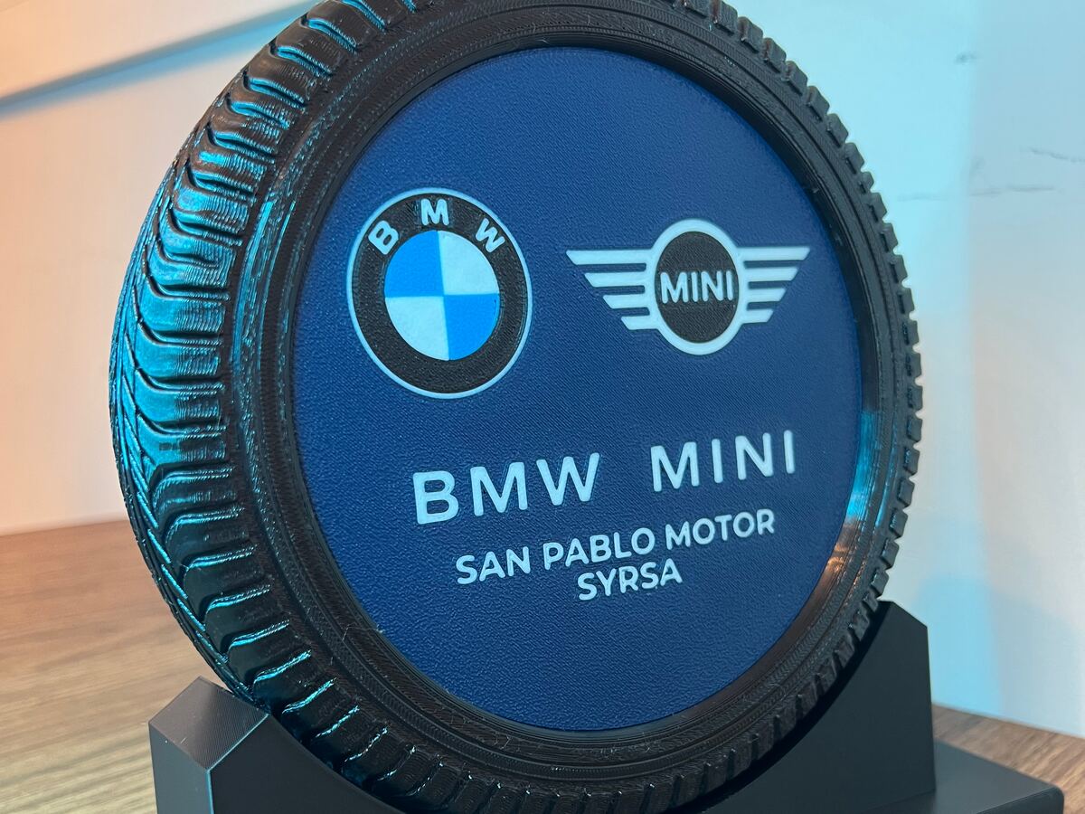 Placa conmemorativa impresa en 3D con forma de neumático, con los logotipos del concesionario en relieve a color sobre un fondo azul y una peana negra